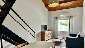 A modern living room with a white table and chairs, a sofa, a TV on a wooden cabinet, and a staircase with black railings.