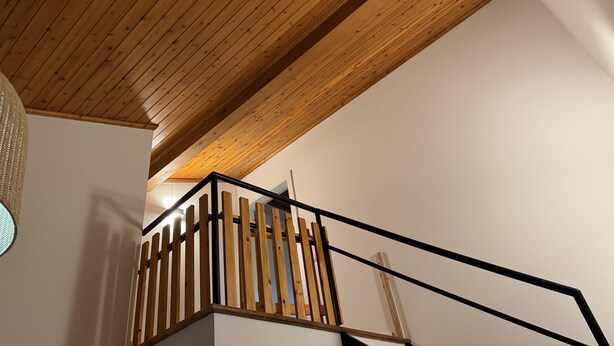 A wooden ceiling with a sloped design, a staircase with wooden railings and black metal handrails, and a white wall.