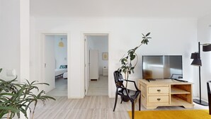 Ein modernes Wohnzimmer mit einem Holztisch, einem Fernseher und einer Pflanze. Ein Durchgang führt in einen anderen Raum.