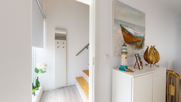 Ein Flur mit weißer Wand, einer Holztreppe, einer kleinen weißen Kommode mit Dekoration und einem Gemälde von Boot und Leuchtturm.