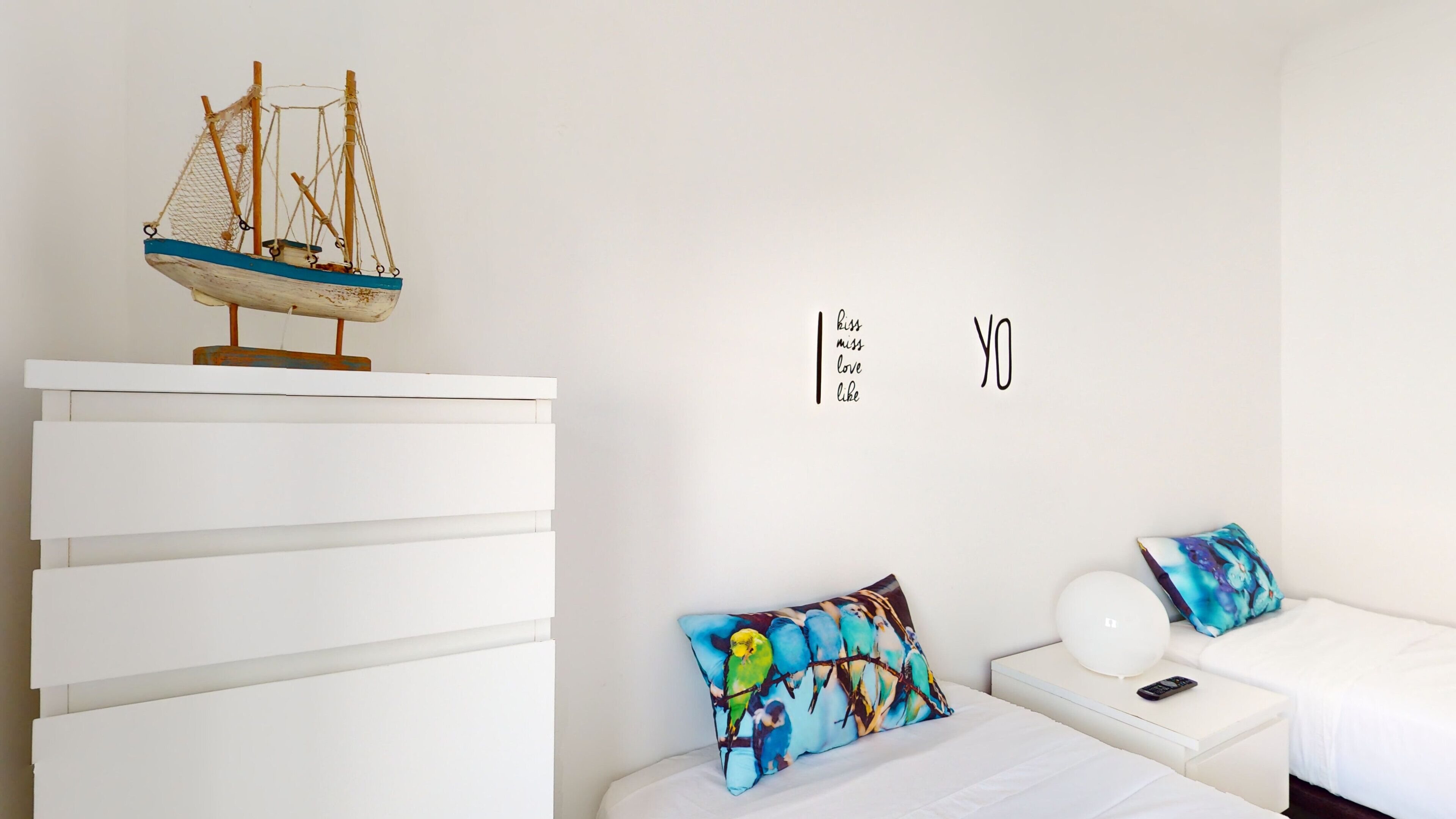 A bedroom with a bed, a nightstand, a dresser, a decorative sailboat, and a wall with the text "kiss miss love like".