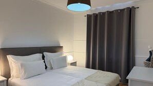 En pænt redt seng med hvide hør og et dekorativt tæppe, en sengetårnslampe og en mørk gardin.