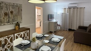 Un coin repas avec une table en verre dressée pour quatre personnes, des chaises blanches et un ananas en guise de décoration centrale. Un luminaire suspendu de couleur marron se trouve au-dessus de la table.