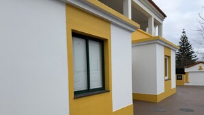 Un bâtiment à l’extérieur jaune et blanc, avec une fenêtre dotée d’un cadre vert et une surface au sol de couleur marron.