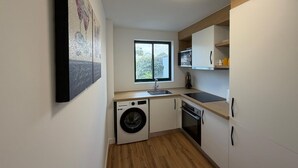 Une cuisine compacte équipée d’un lave-linge, d’un four et d’une fenêtre donnant sur un espace verdoyant.