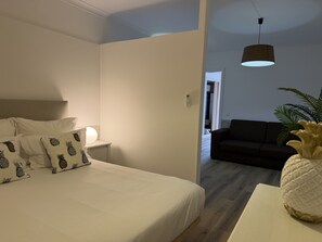 Un dormitorio con cama, mesita de noche y lámpara. Se ve un pasillo que conduce a una sala de estar con sofá y una planta.