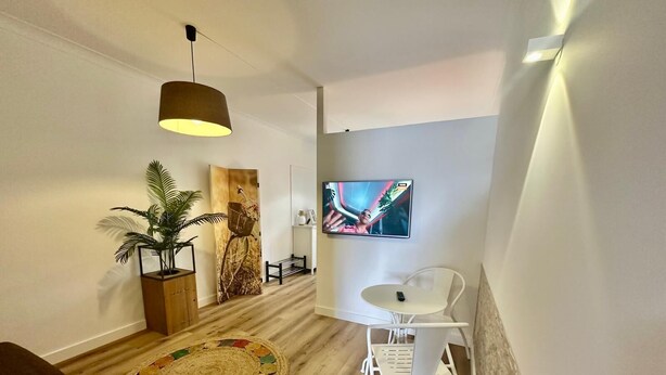 Un salon moderne avec un plancher en bois, un tapis rond en fibres naturelles, une plante en pot, une chaise blanche, une petite table, un téléviseur fixé au mur et un luminaire suspendu.