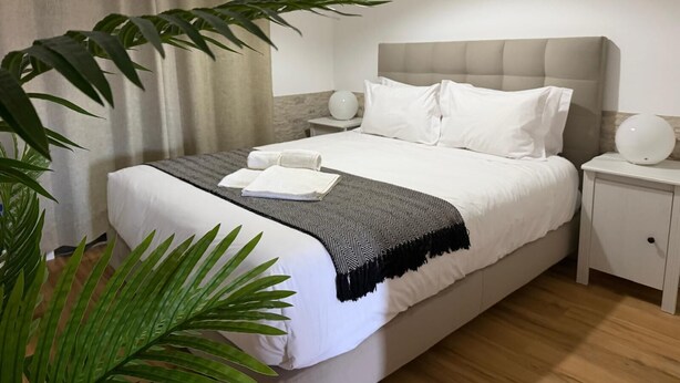 A neatly made bed with white linens, a grey and black patterned throw blanket, and two white towels. There are two bedside tables with white lamps, and a large green plant is visible to the left.
