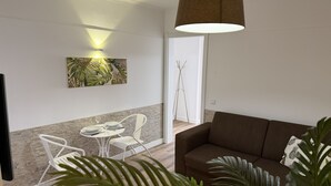 A modern living room with a brown sofa, a white dining table with chairs, a large plant, and a wall-mounted lamp.