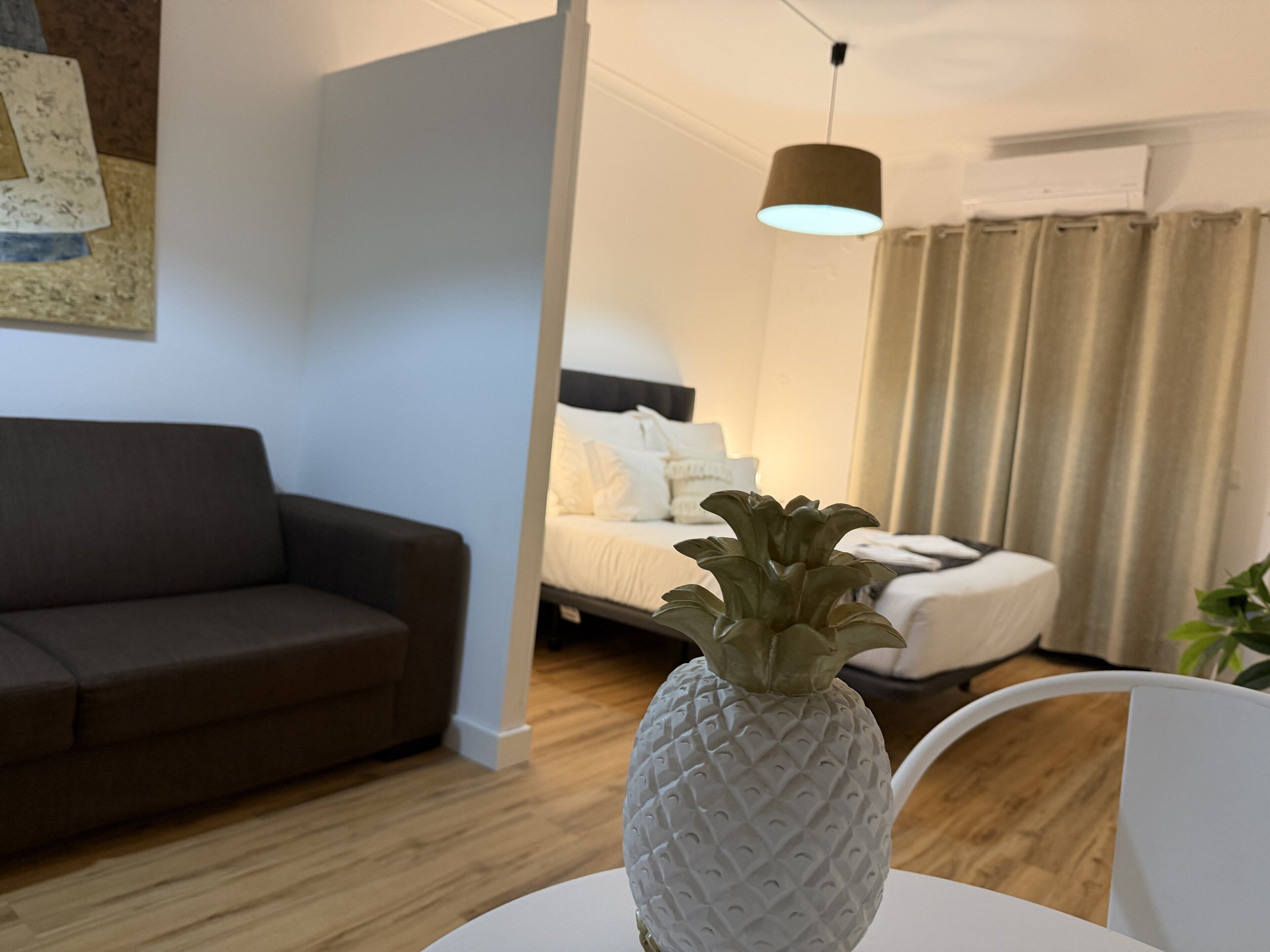 Ett modernt vardagsrum med en soffa, en säng med vita sängkläder, en vas formad som en ananas och en hängande lampa.