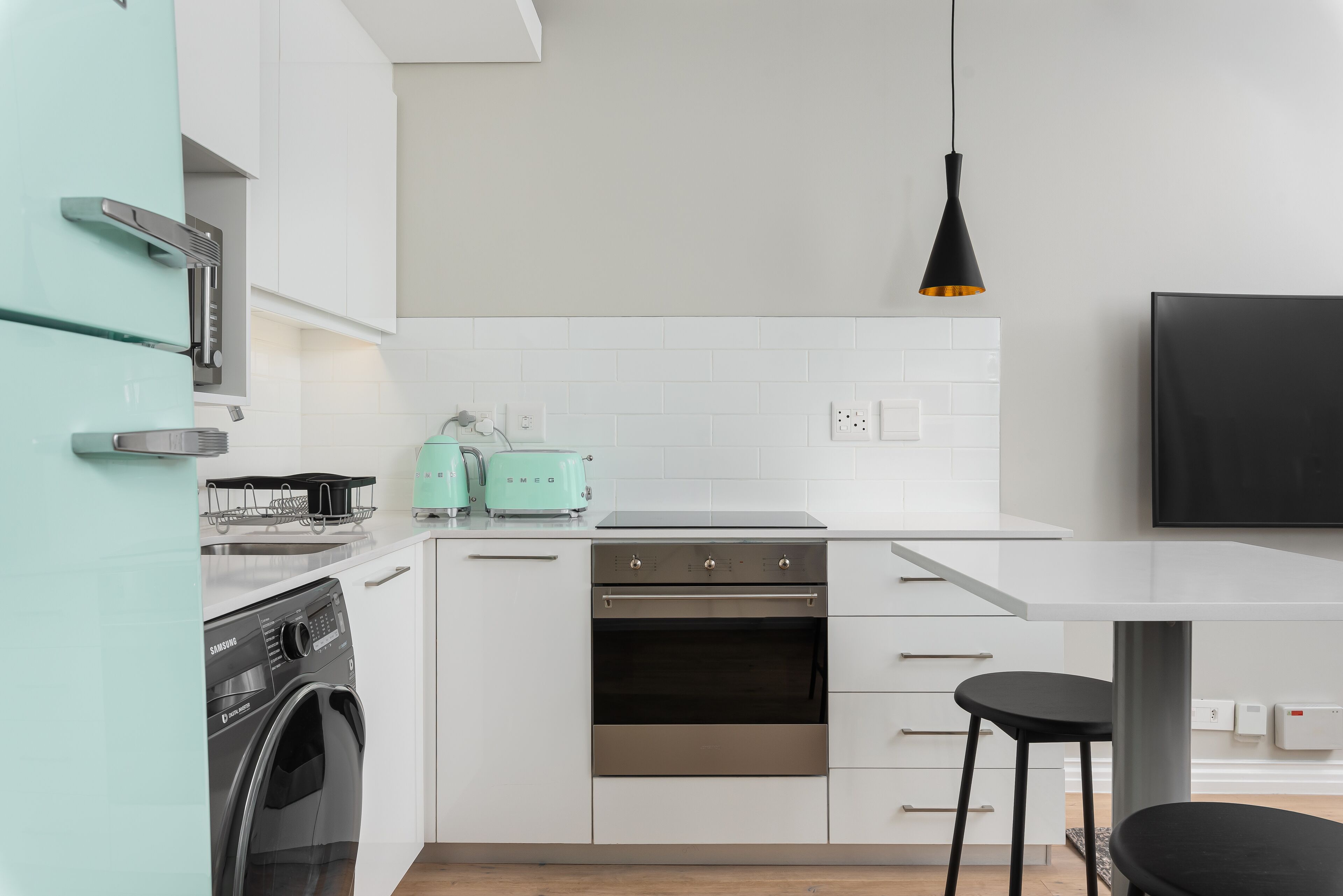 Une cuisine moderne avec des meubles blancs, un four en acier inoxydable, un lave-linge, un grille-pain et un luminaire suspendu.