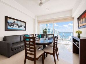 A modern living room with a dining table, chairs, a sofa, and a balcony with a view of the sea and buildings.