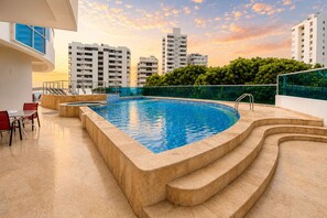 A rooftop swimming pool with steps leading into the water, surrounded by a glass railing and a view of high-rise buildings.