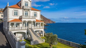 Un grand bâtiment blanc, surmonté d’un toit recouvert de tuiles rouges, doté d’un balcon et d’un escalier menant à l’entrée. Il offre une vue sur une étendue d’eau et un paysage montagneux.