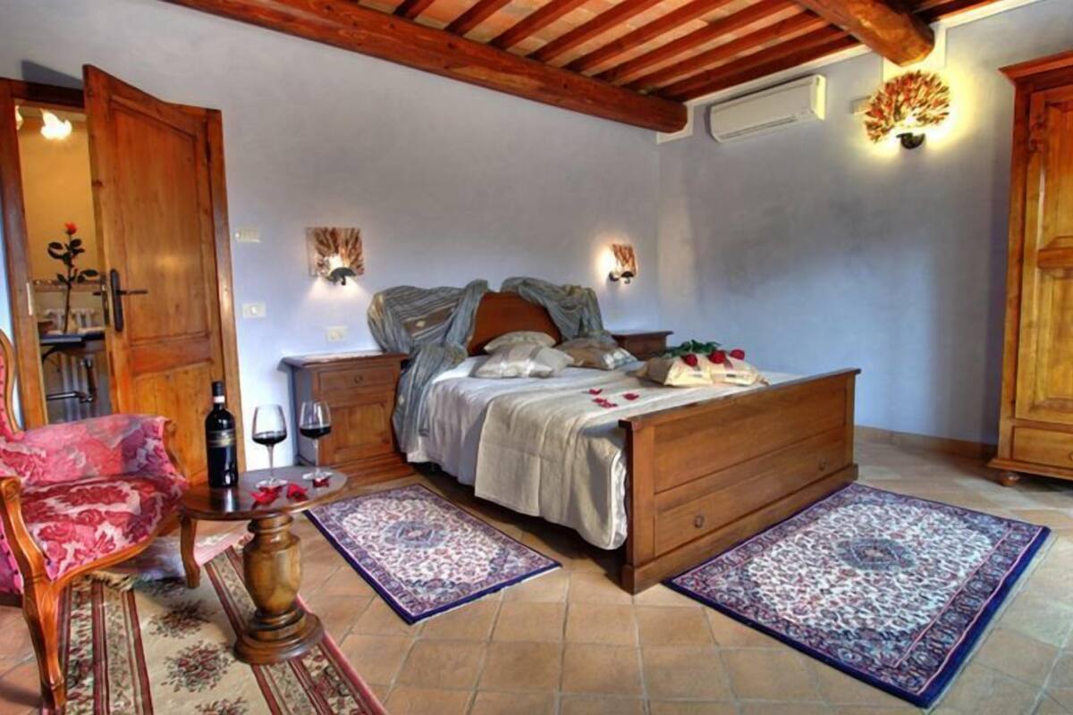 Quarto com cama de madeira, uma cadeira vermelha, mesa de cabeceira com garrafa e copos, e uma planta verde grande.