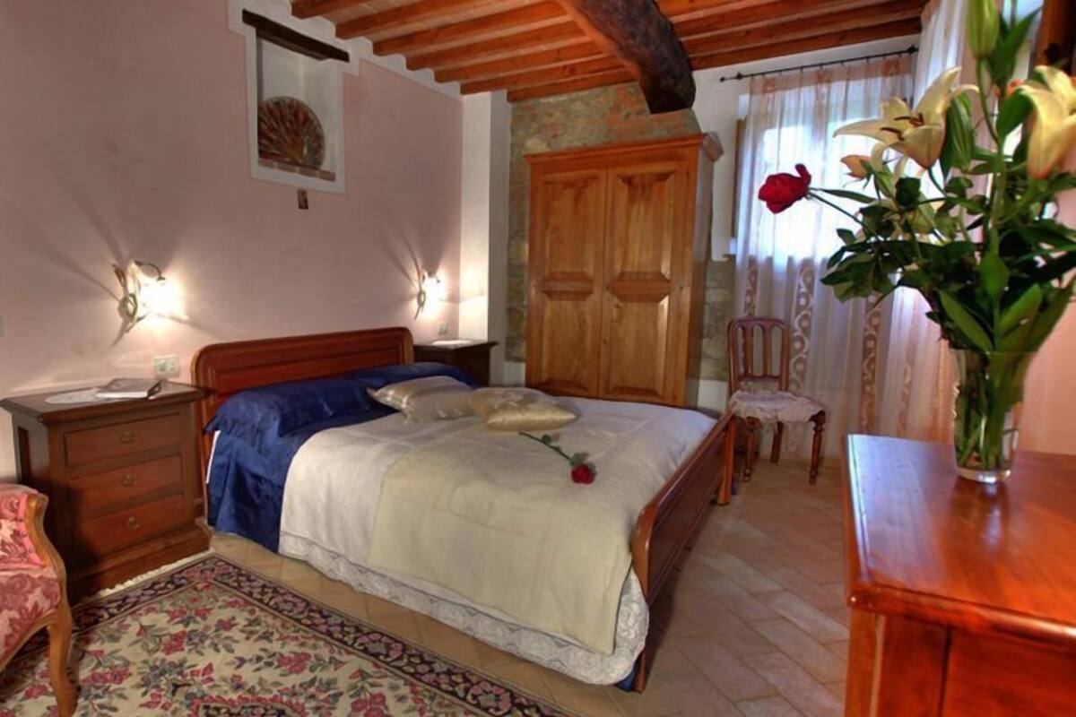 Quarto com cama de madeira, mesa de cabeceira, cômoda, cadeira e um vaso com flores.