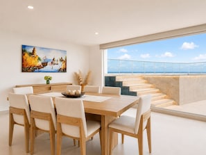 A modern dining area with a wooden table and chairs, a painting on the wall, and a view of the sea through large windows.