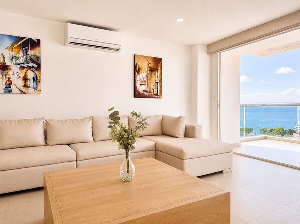 A modern living room with a beige sofa, a wooden coffee table, and a vase with green plants. There are two paintings on the wall, and an air conditioning unit is mounted above the sofa. - Penthouse • Private pool • Ocean view • Beach 1803 (Cartagena)