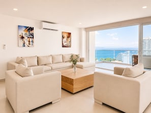 A modern living room with a beige sofa, two armchairs, a wooden coffee table, and a large window with a view of the sea and buildings. - Penthouse • Private pool • Ocean view • Beach 1803 (Cartagena)
