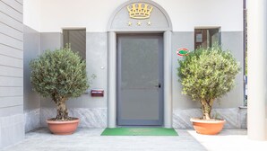 Entrada de un edificio con una puerta gris, un emblema con forma de corona y dos árboles en macetas.
