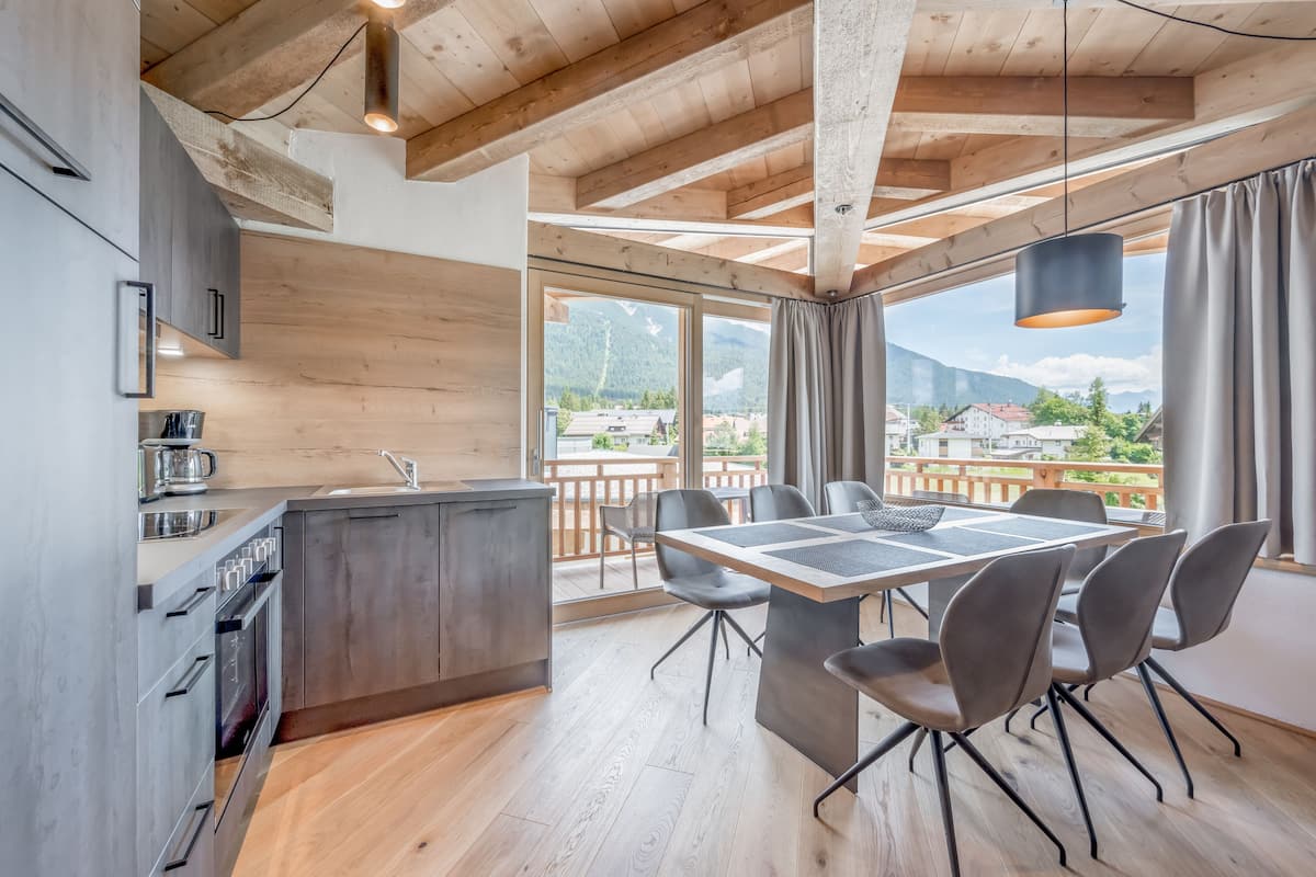 A modern kitchen with wooden beams, a dining area with a table and chairs, and a large window with a view of mountains.