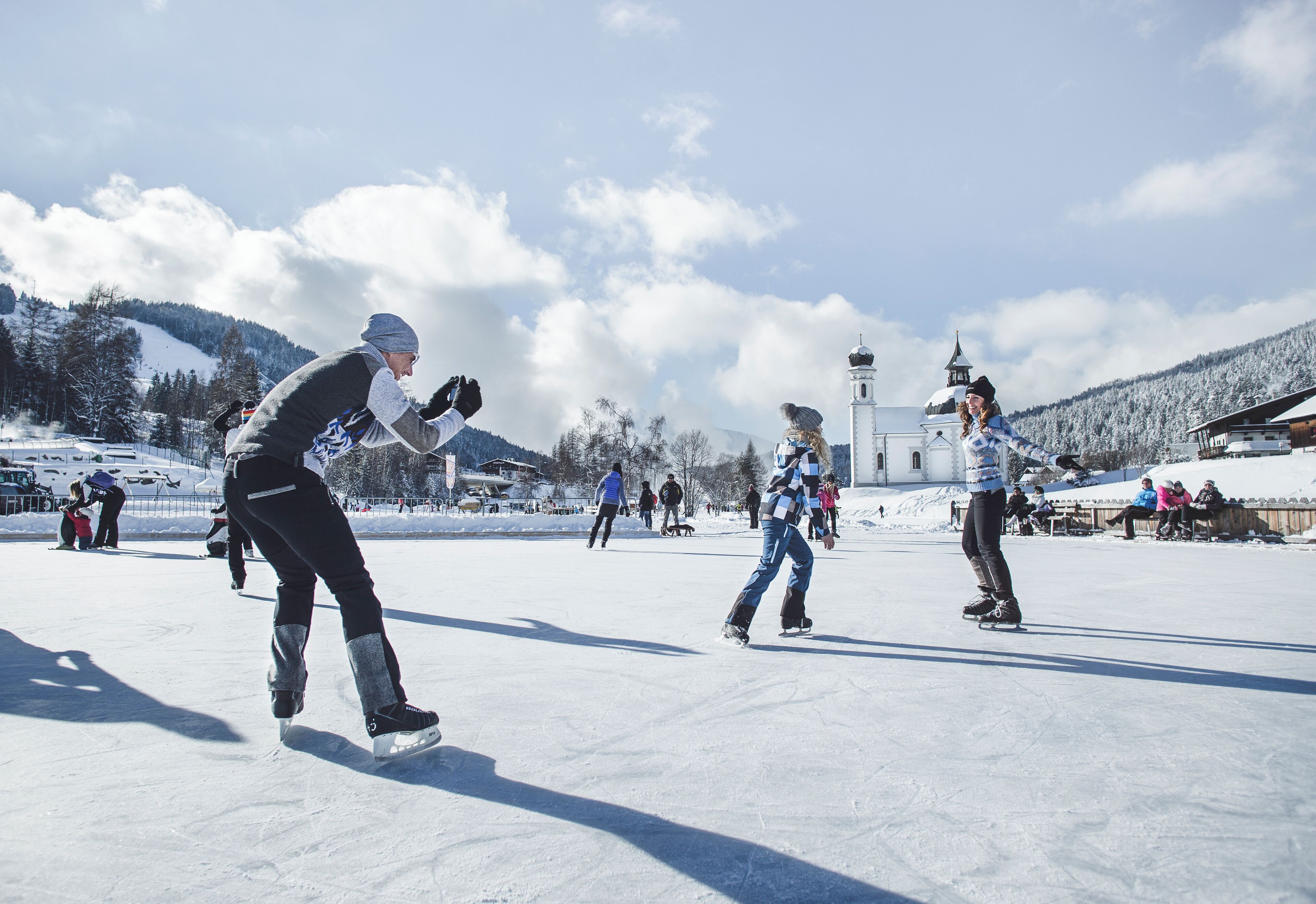A group of people ice skating on a snow-covered surface with a church and mountains in the background.