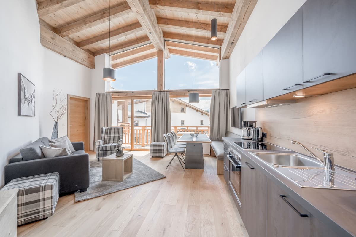 A modern kitchen with a wooden ceiling, a dining area with a table and chairs, and a living room with a sofa and ottoman.