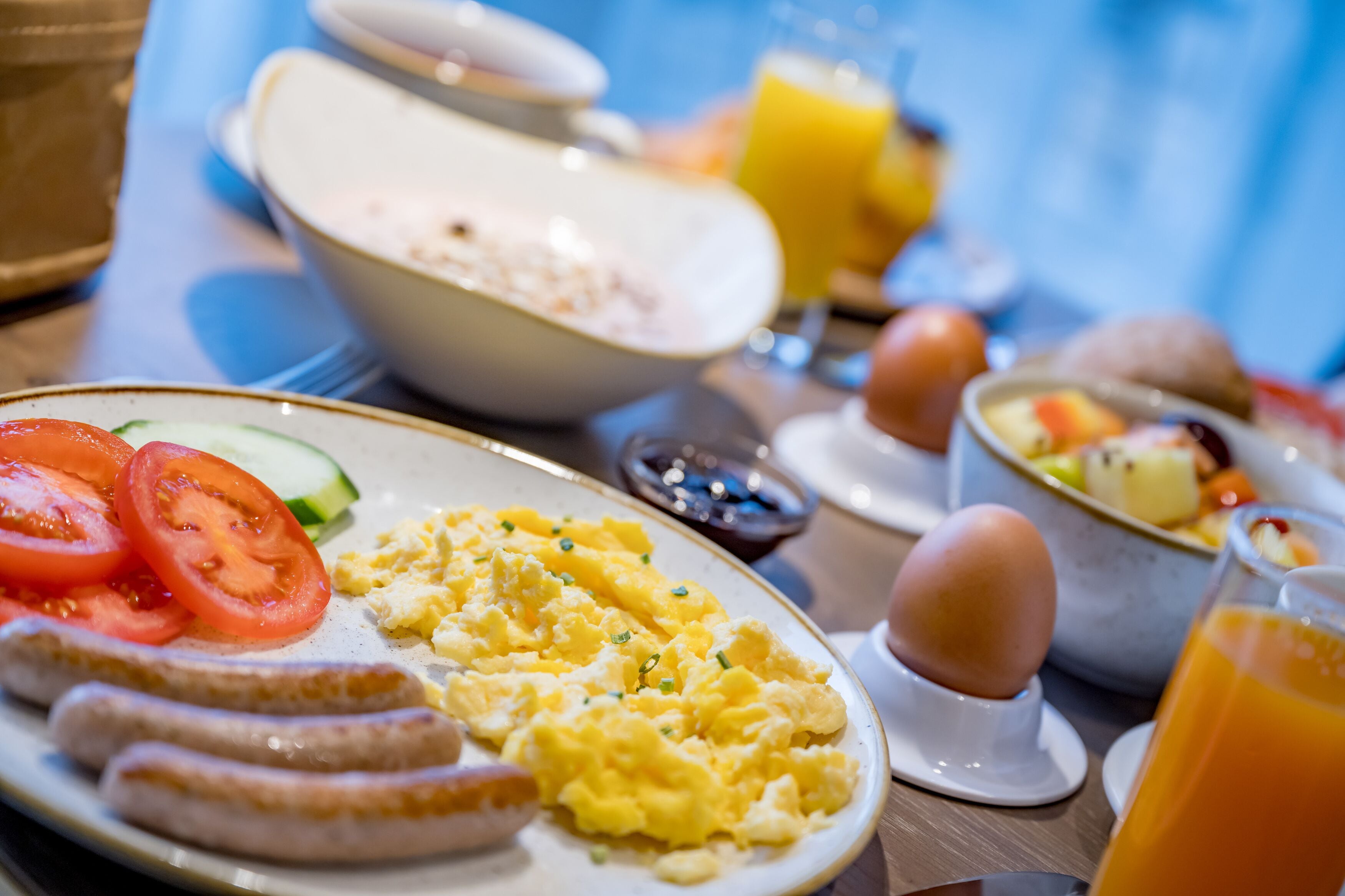 Una mesa de desayuno con huevos revueltos, salchichas, tomates y un tazón de cereal.