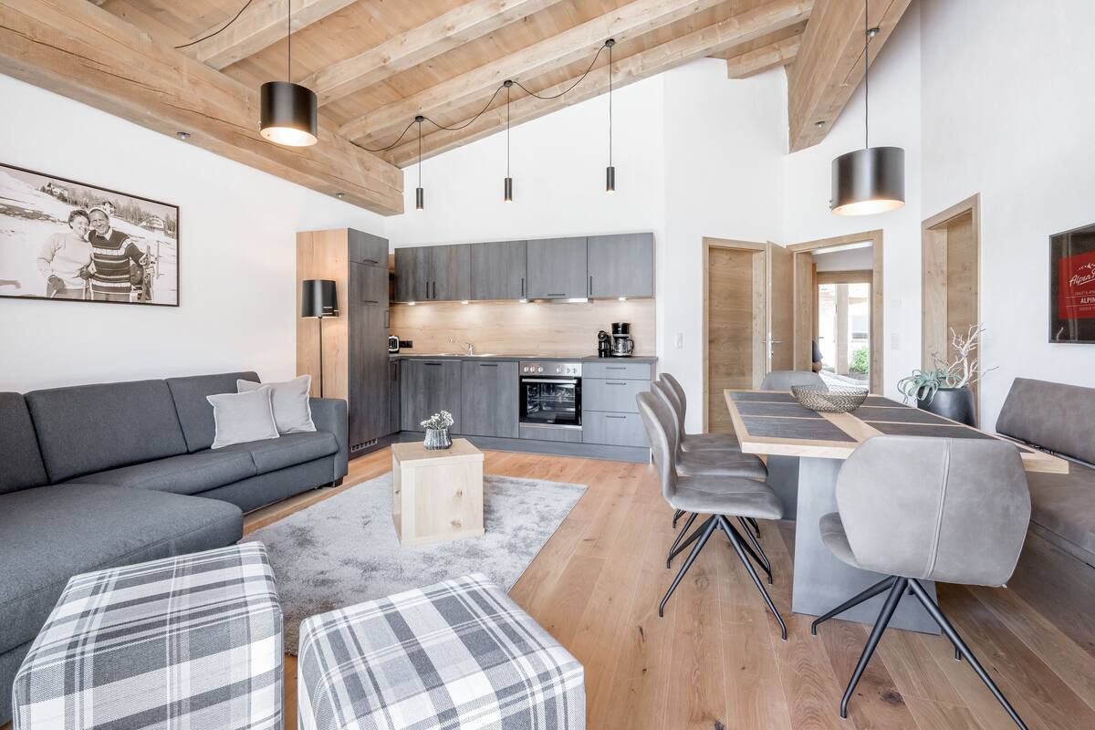 A modern living space with a grey sofa, a dining table with chairs, a kitchen area, and a wooden ceiling.