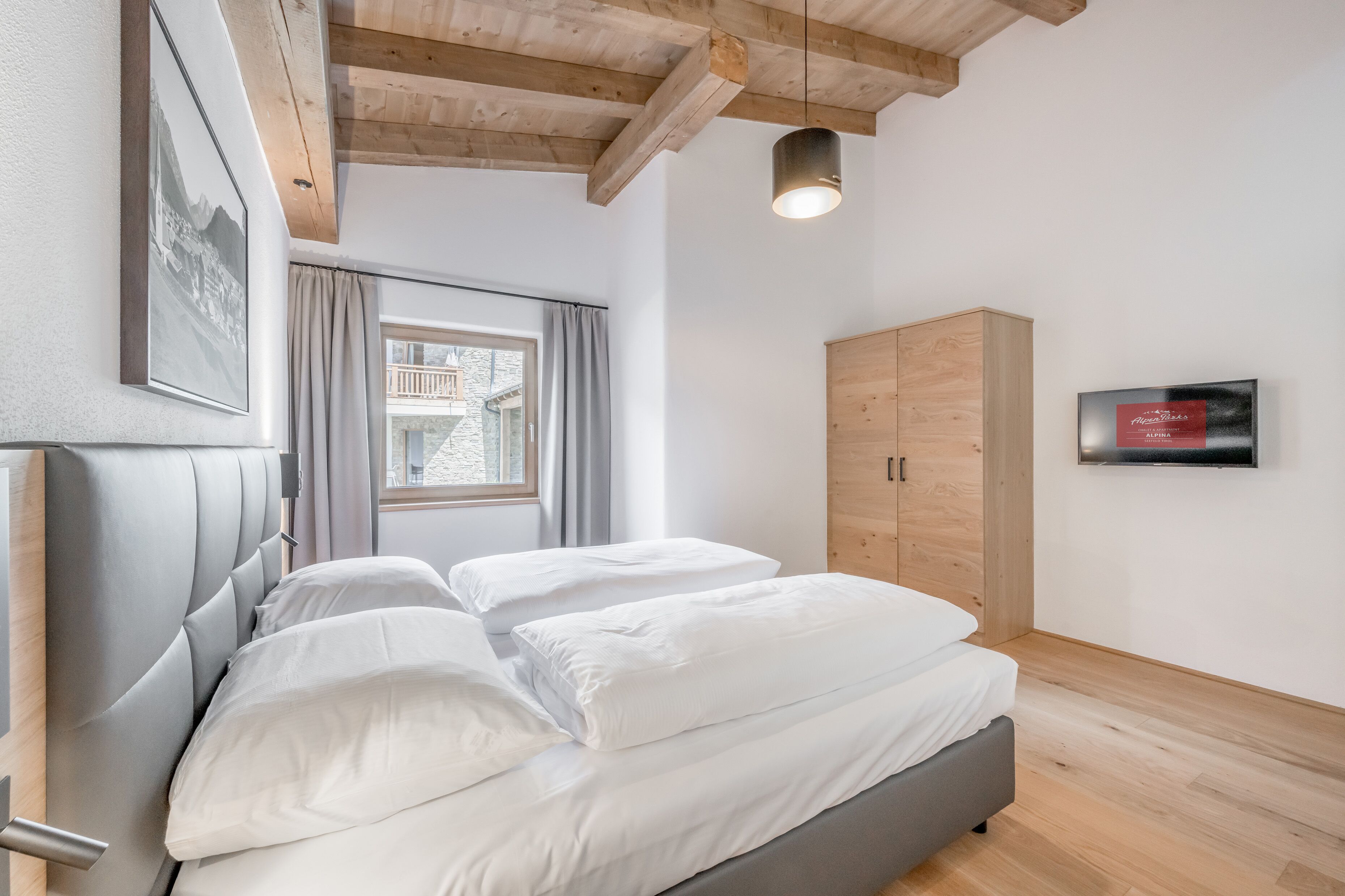 Un dormitorio moderno con cama, vigas de madera en el techo, una ventana con cortinas, un armario y un televisor de pared.