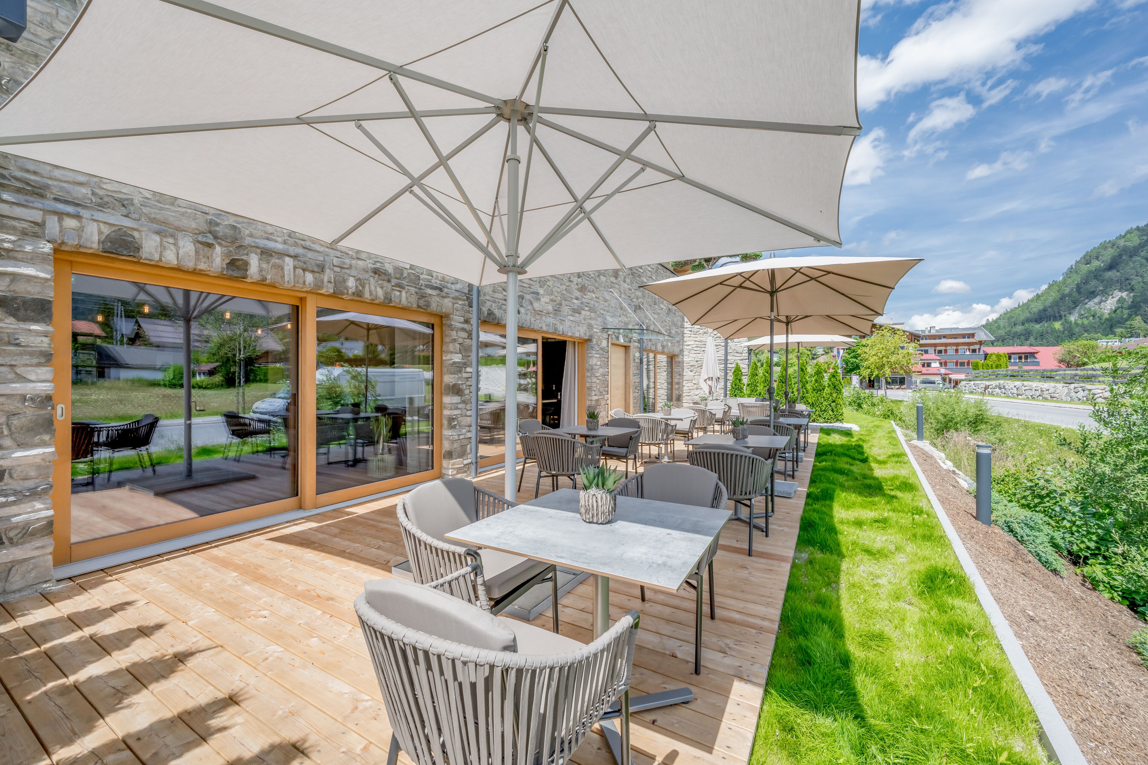 En udendørs terrasse med borde og stole, store parasoller og en bygning i sten med glasdøre.