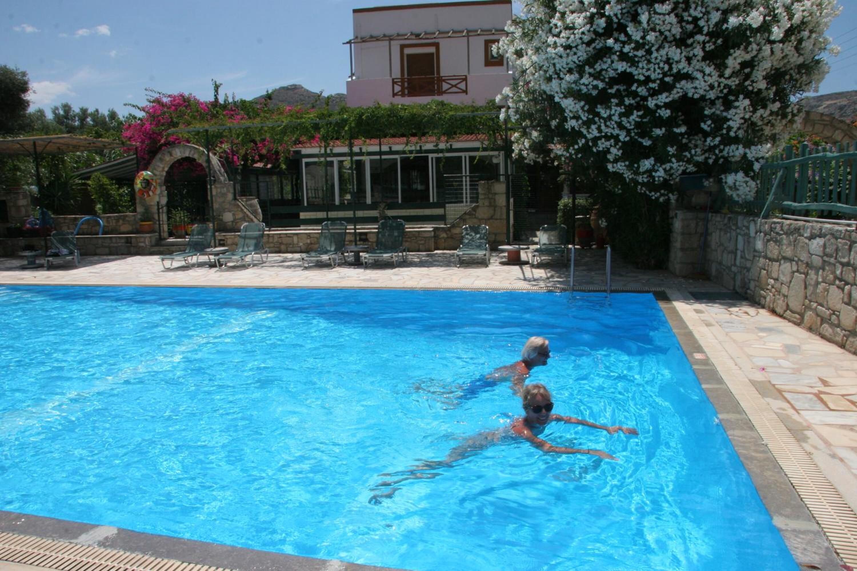 A swimming pool with two people in the water, surrounded by stone walls and a building with a balcony.