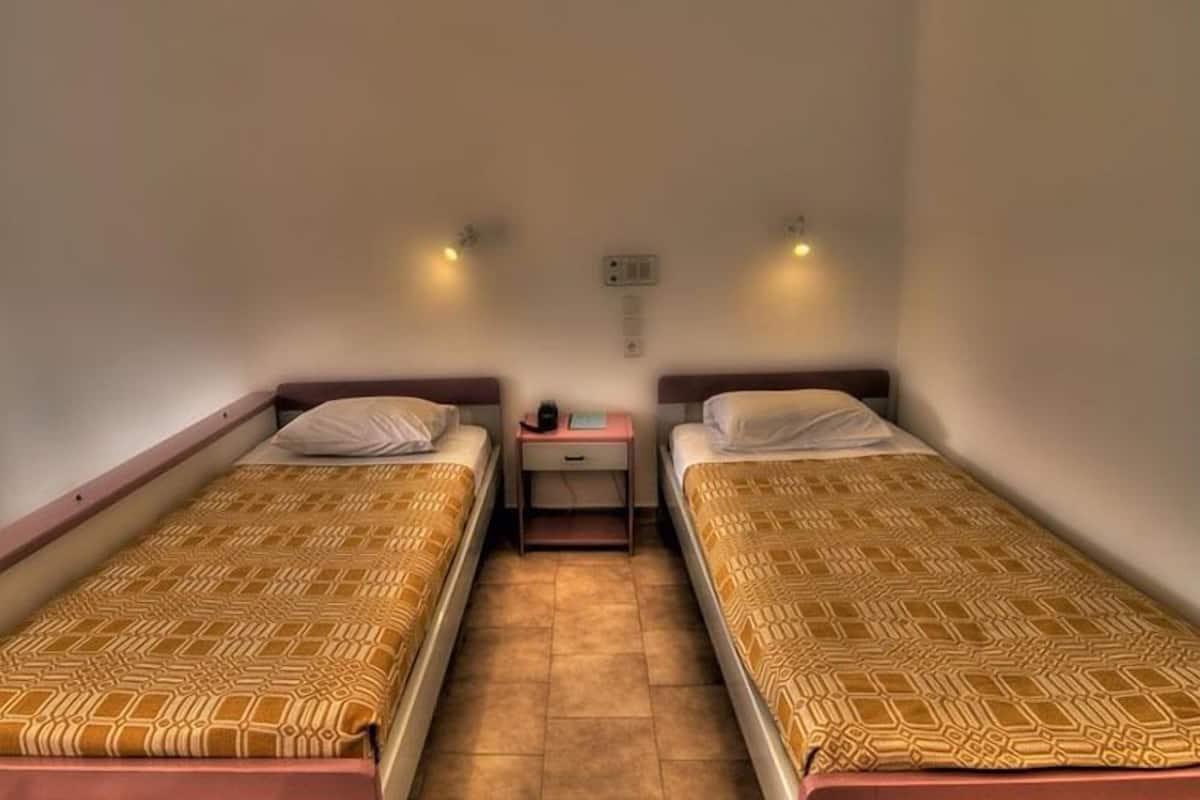 Acomodação modesta, com um quarto individual, duas camas de solteiro, uma mesinha de cabeceira pequena e luminária fixada na parede.
