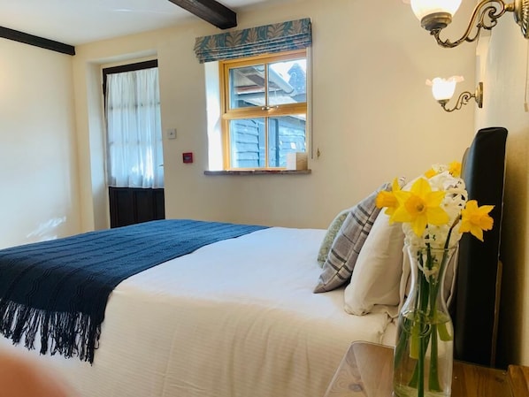 Ein Schlafzimmer mit Bett, einem Fenster mit Vorhängen, einer Vase mit Blumen und einer Lampe.