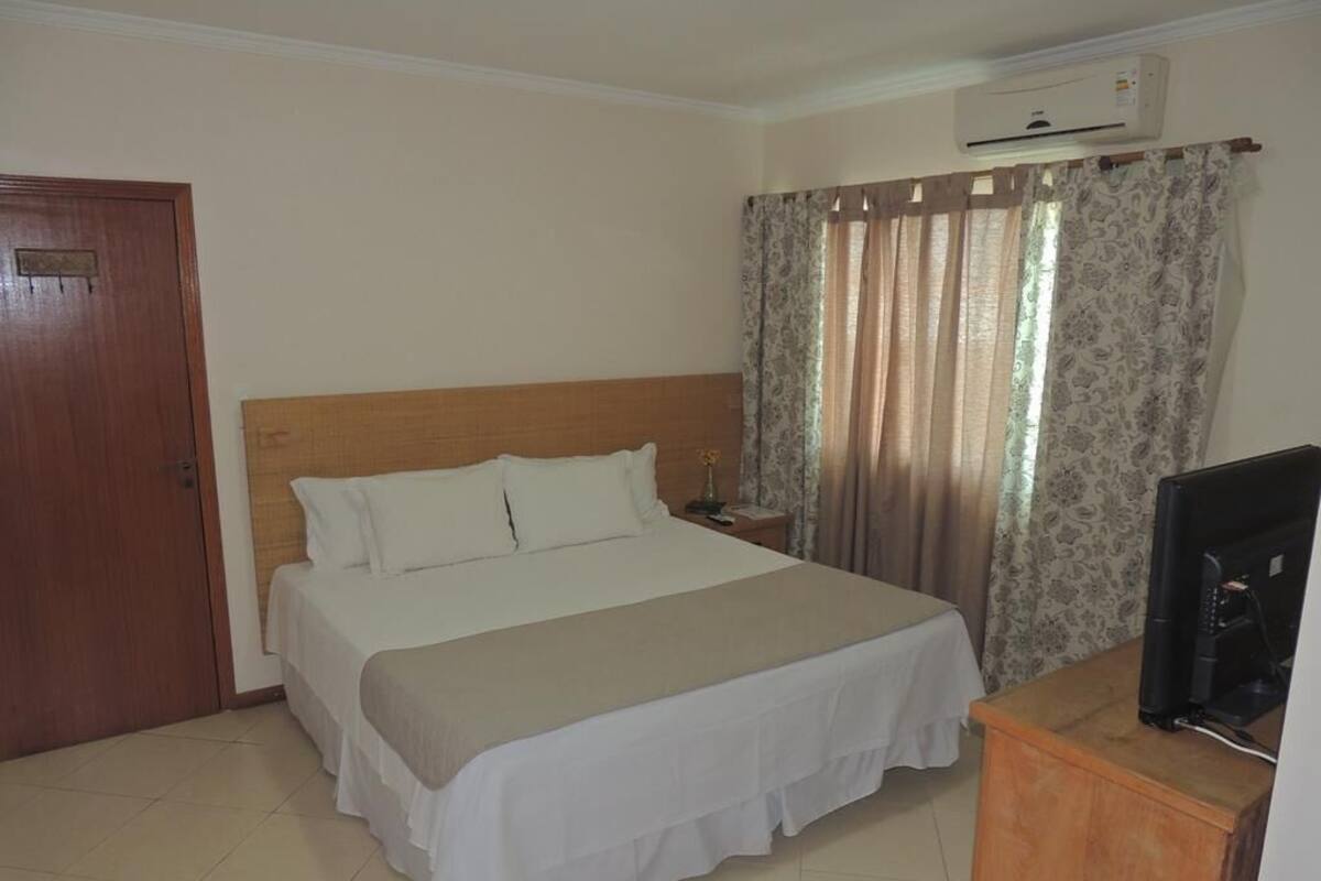 A hotel room with a bed, a wooden headboard, a nightstand, a TV, and a window with curtains.