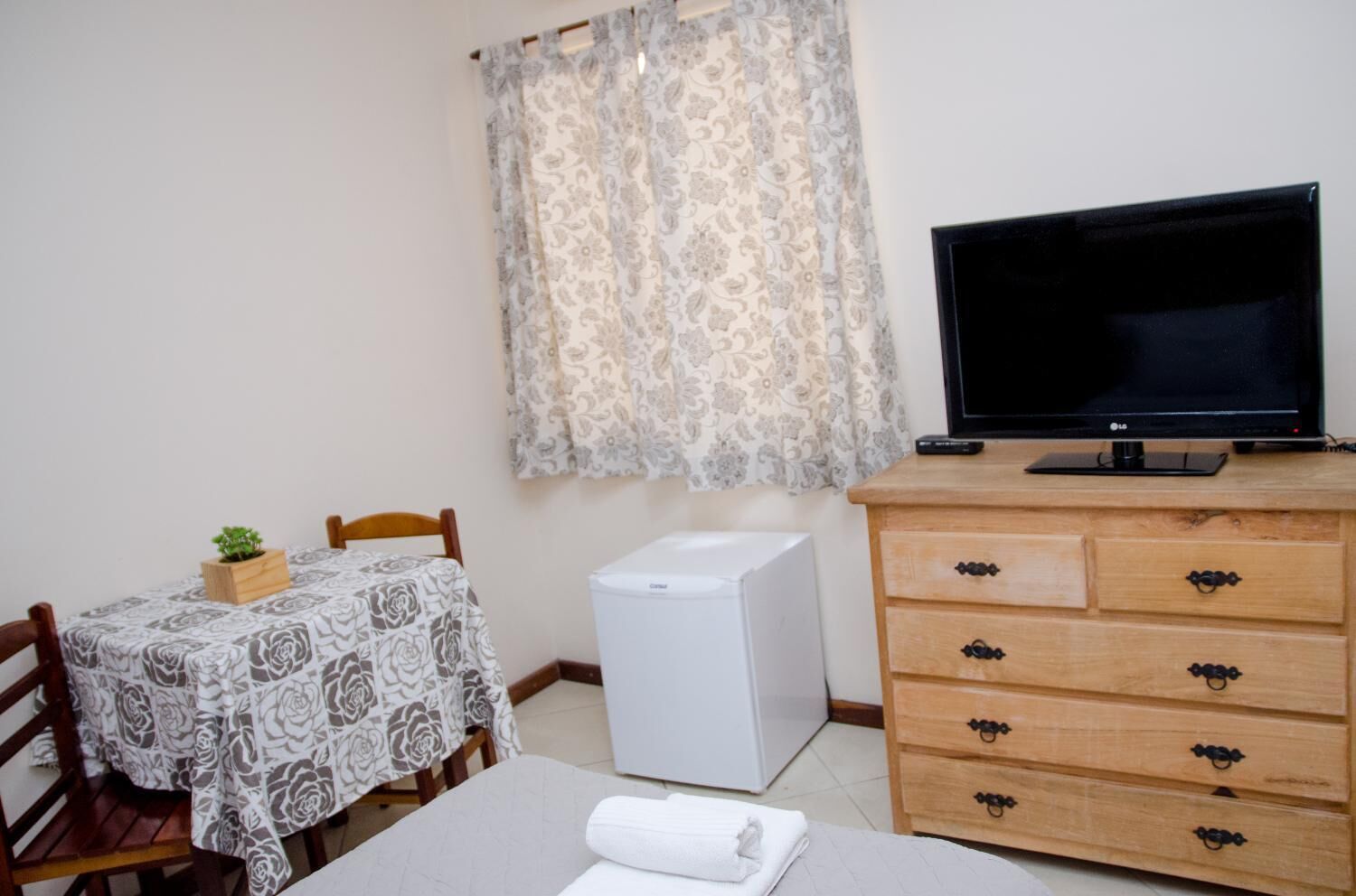 Quarto pequeno com uma mesa de jantar, cadeiras, uma TV sobre uma cômoda de madeira, um radiador branco e uma cortina estampada.