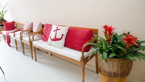 Une rangée de bancs en bois, dotés de coussins blancs et d’oreillers rouges, chacun présentant un motif différent. Des plantes en pot, ornées de fleurs rouges, sont disposées sur le côté droit.