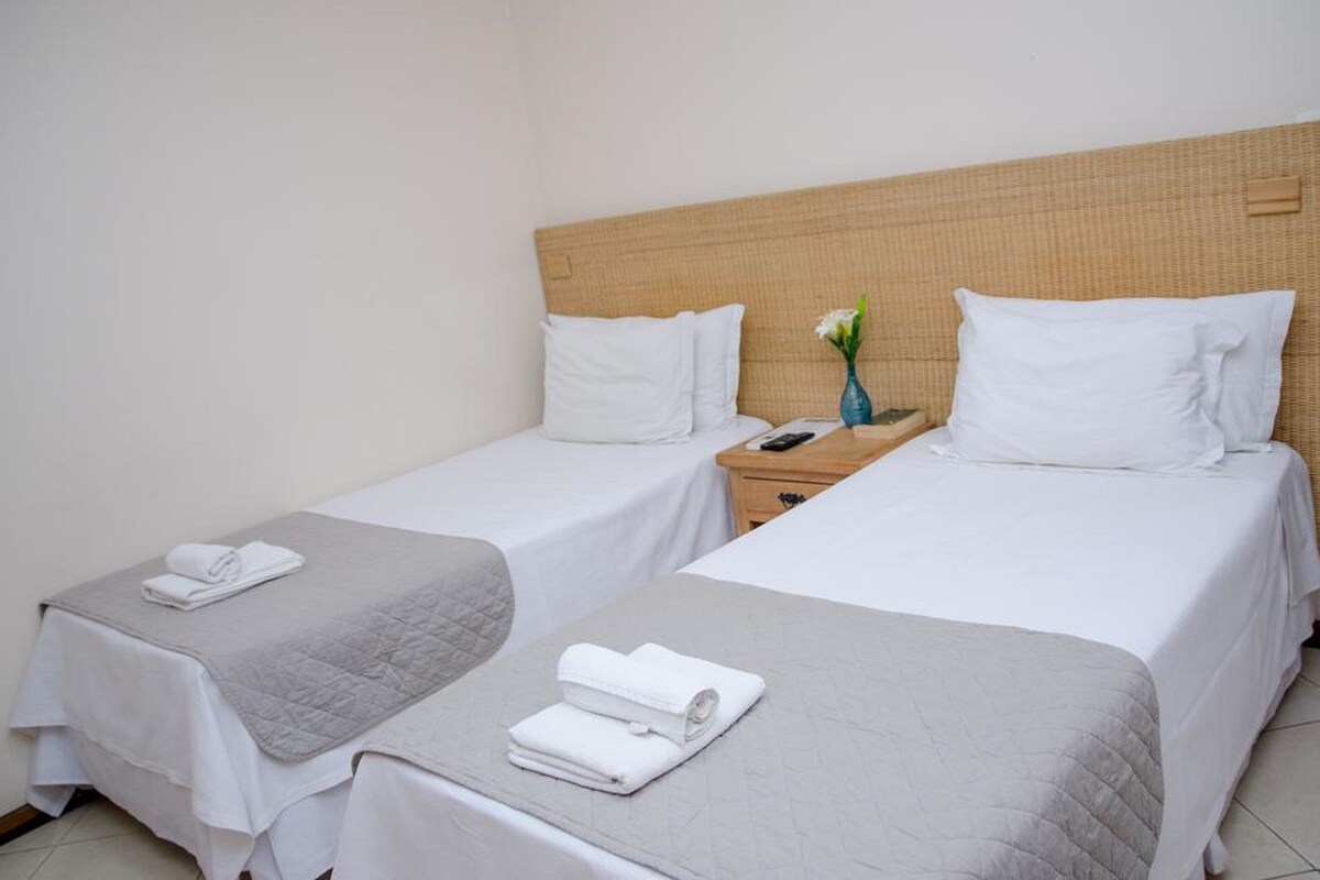 Habitación de hotel con dos camas individuales, cada una con una colcha blanca y un edredón gris. Hay una mesita de noche de madera entre las camas, con un jarrón de flores y un control remoto.
