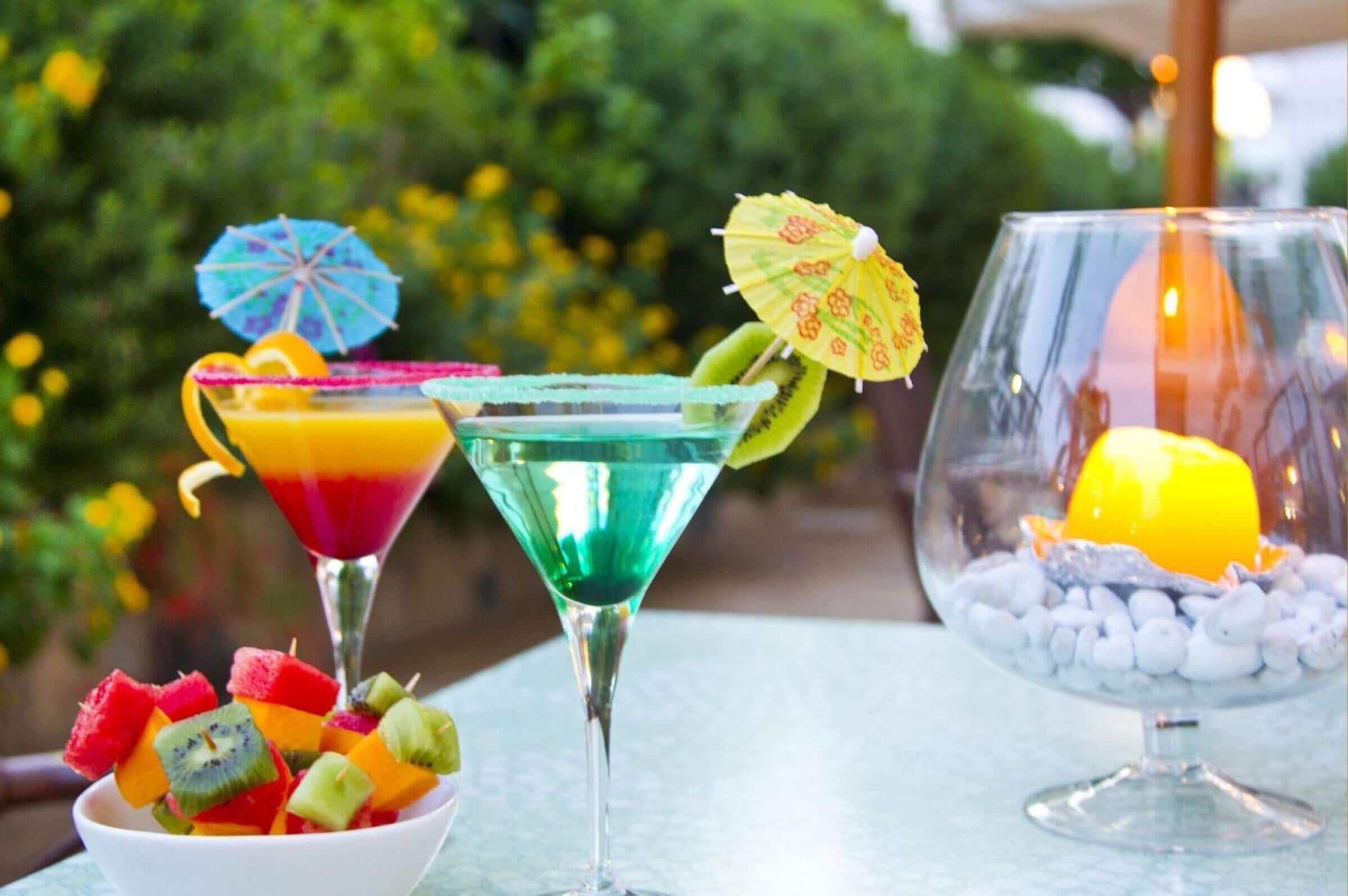 Tre cocktail con guarnizioni di frutta e ombrellini, una ciotola di frutta e una candela in un bicchiere.