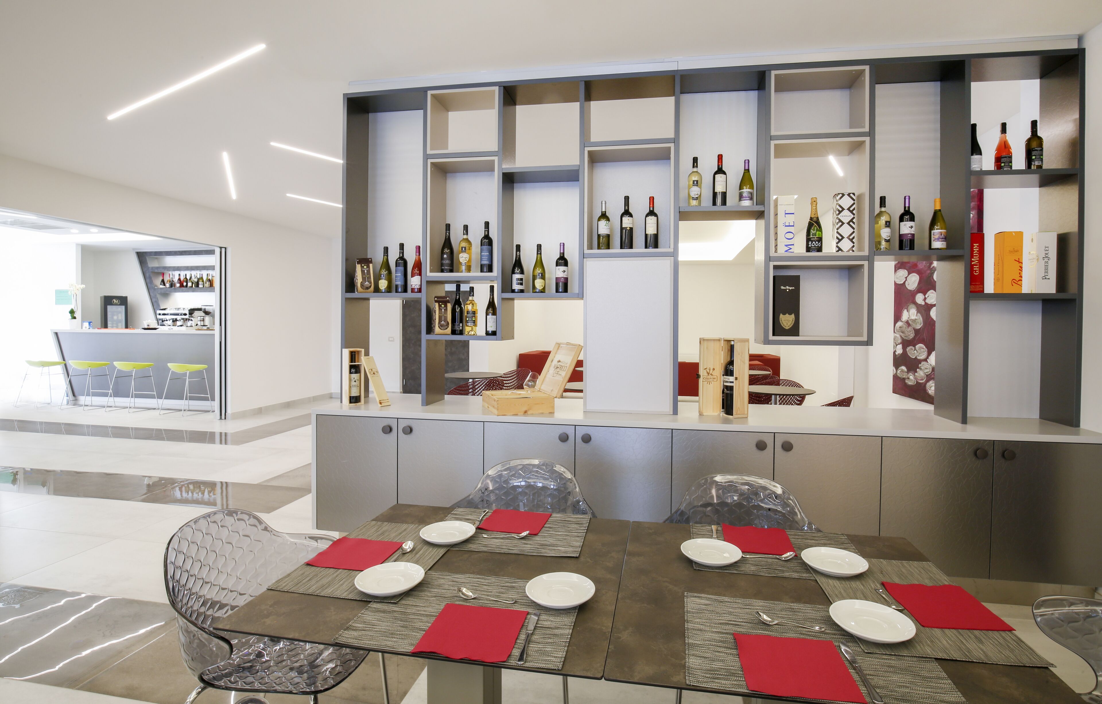 Un comedor moderno con una mesa de vidrio dispuesta para cuatro personas, rodeada de sillas metálicas. Un mostrador de vinos se encuentra detrás de la mesa, y se distingue una zona de bar al fondo.