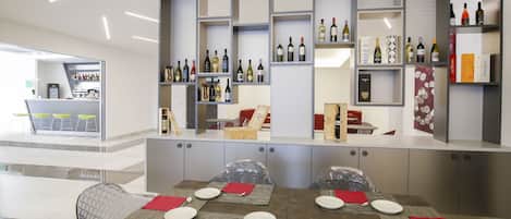 Un comedor moderno con una mesa de vidrio dispuesta para cuatro personas, rodeada de sillas metálicas. Un mostrador de vinos se encuentra detrás de la mesa, y se distingue una zona de bar al fondo.
