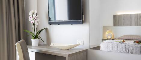 Habitació d'hotel moderna amb televisió de pantalla plana penjada a la paret, escriptori de fusta amb un lavabo blanc, orquídia en test, llit amb edredó blanc i tauleta de nit amb una làmpada.