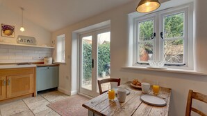 Une cuisine avec une table en bois mise pour le petit-déjeuner, une fenêtre donnant sur un paysage verdoyant et une porte menant à l’extérieur.