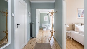 A hallway with a wooden floor, a coat rack, a radiator, and a bed with white bedding in the adjacent room. - Scandinavian Apartments Fredrikinkatu (Helsinki)