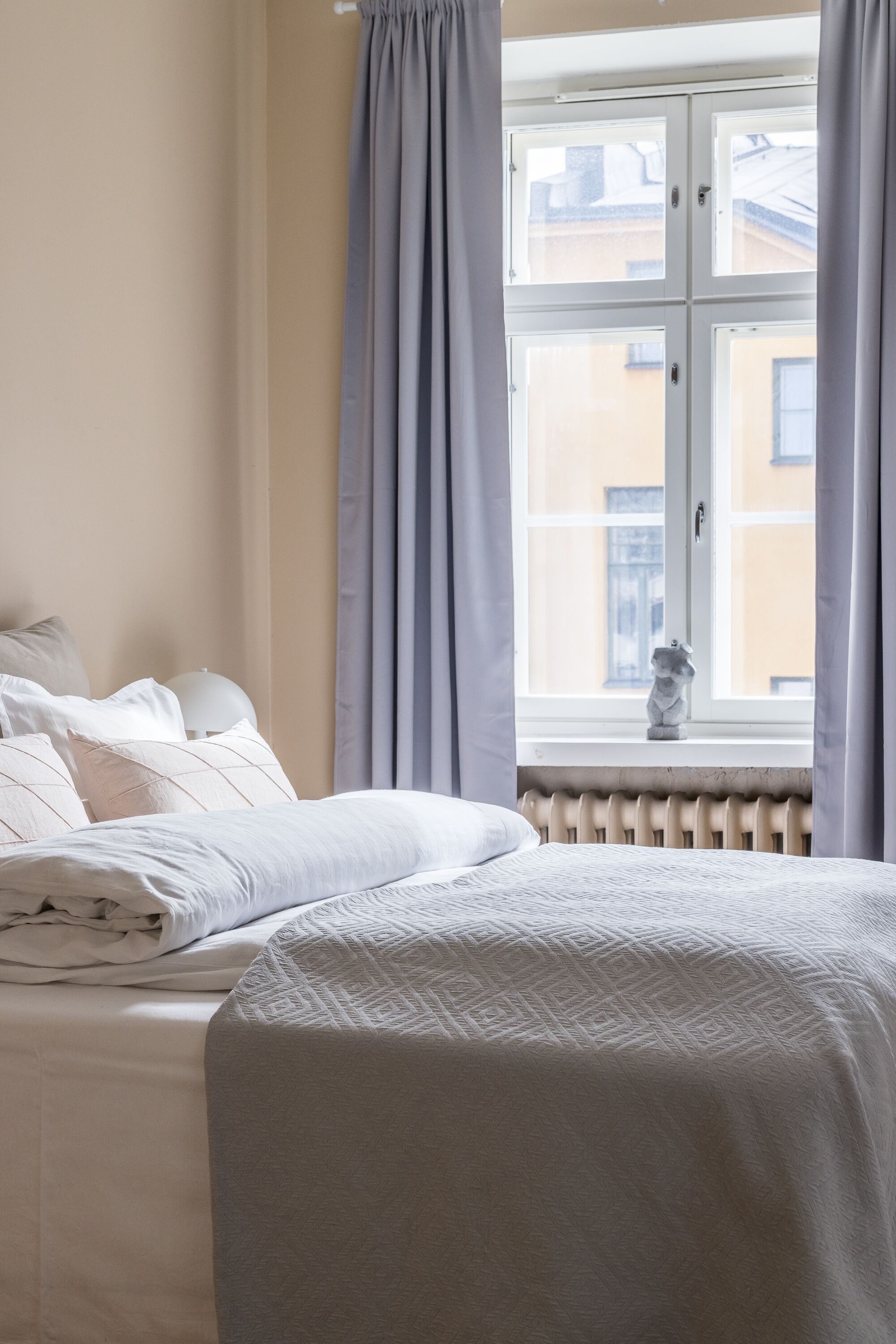 A neatly made bed with a white comforter and pillows, a window with curtains, and a radiator below.
