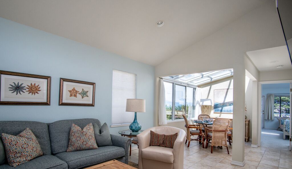 A living room with a sofa, armchairs, a coffee table, and a dining area with chairs and a table. There are framed pictures on the wall, a lamp on a side table, and a window with a view.