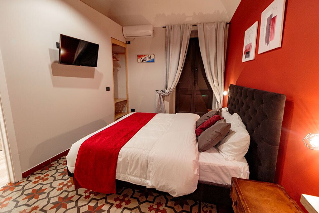 Una camera d'albergo con un letto, una televisione fissata al muro, una parete di colore rosso e un pavimento decorato.