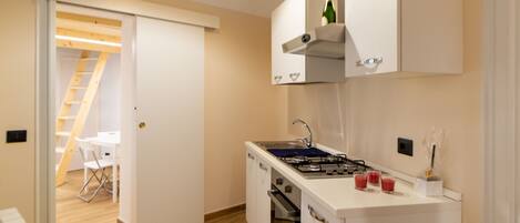 Una cocina compacta con estufa a gas, horno y armarios blancos. Se observa, al fondo, un pequeño comedor con una mesa y sillas.