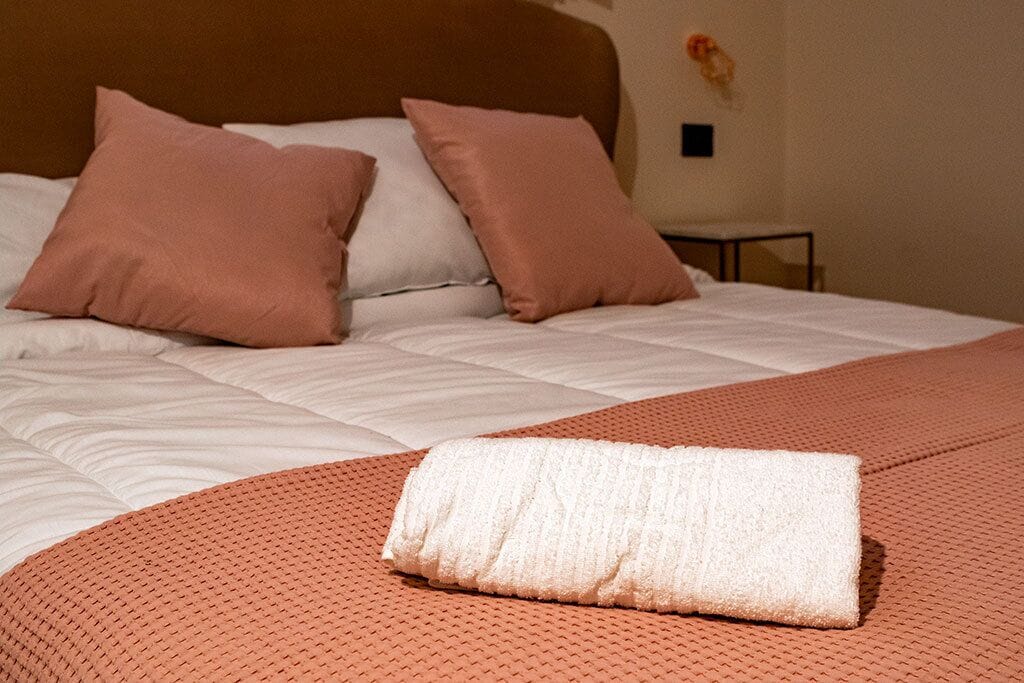 A neatly made bed with a white duvet, two pink pillows, and a folded white towel on an orange textured bedspread.