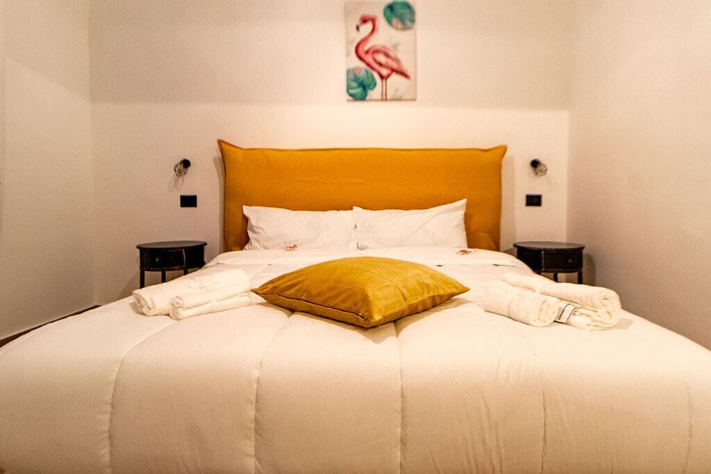Un lit bien fait, recouvert d’une couette blanche et agrémenté d’un coussin de couleur moutarde, flanqué de deux tables de chevet noires sur lesquelles sont posées des lampes.